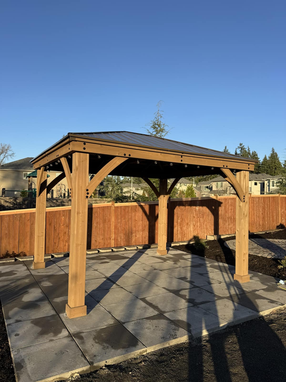 Cedar gazebo with dark metal roof on a paver patio under clear blue sky
