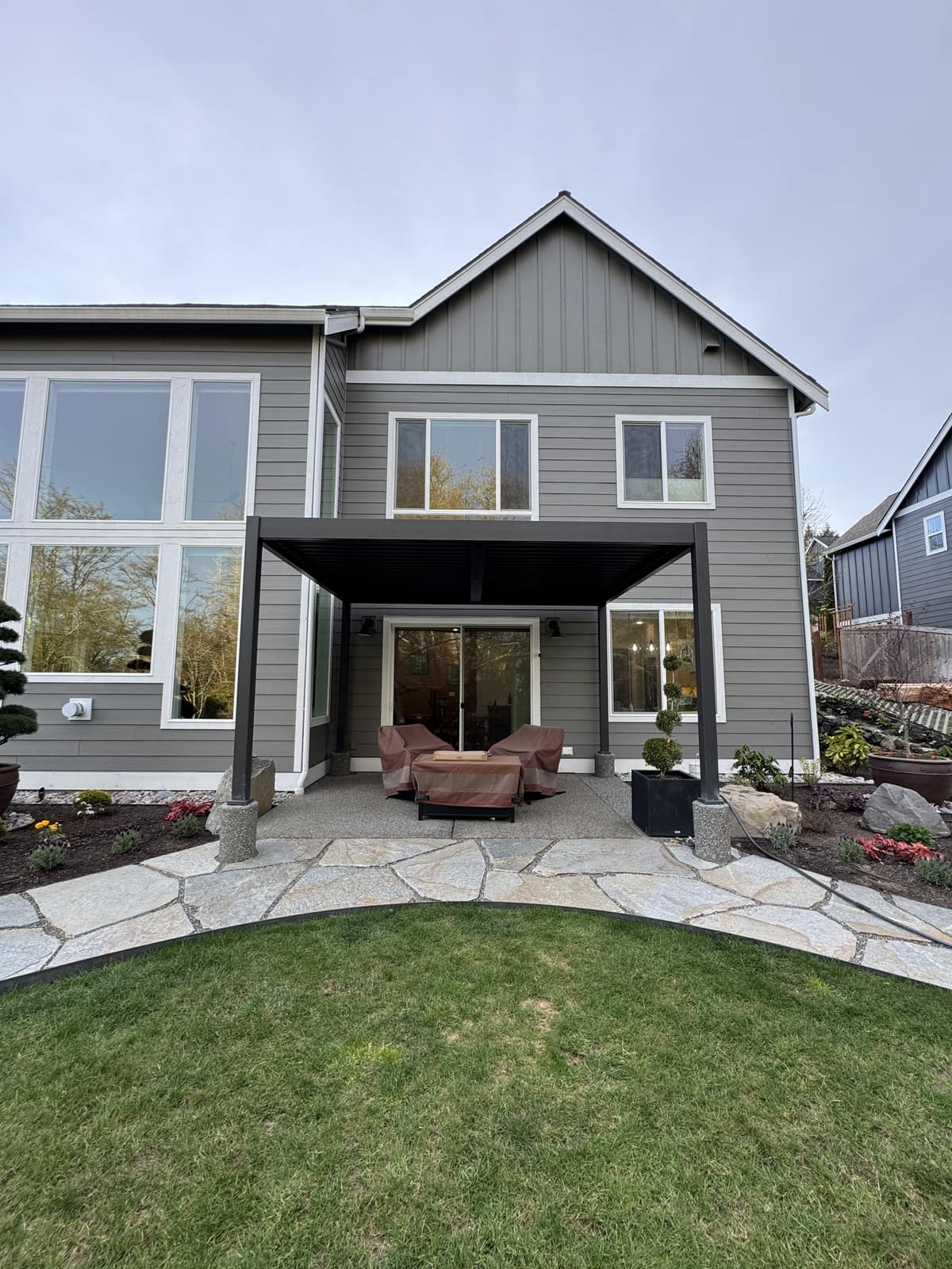Black louvered pergola attached to a modern gray home over a flagstone patio