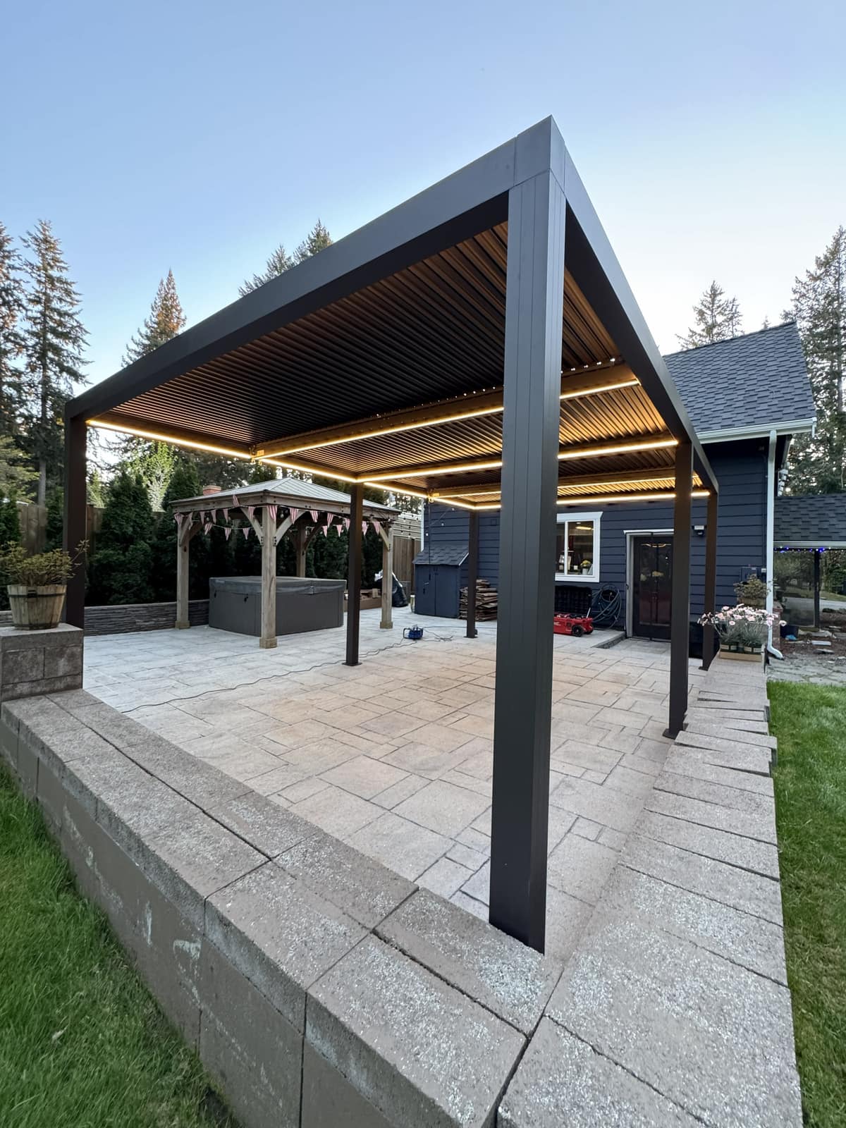 Black louvered pergola glowing with warm LED lighting at dusk on a raised paver patio