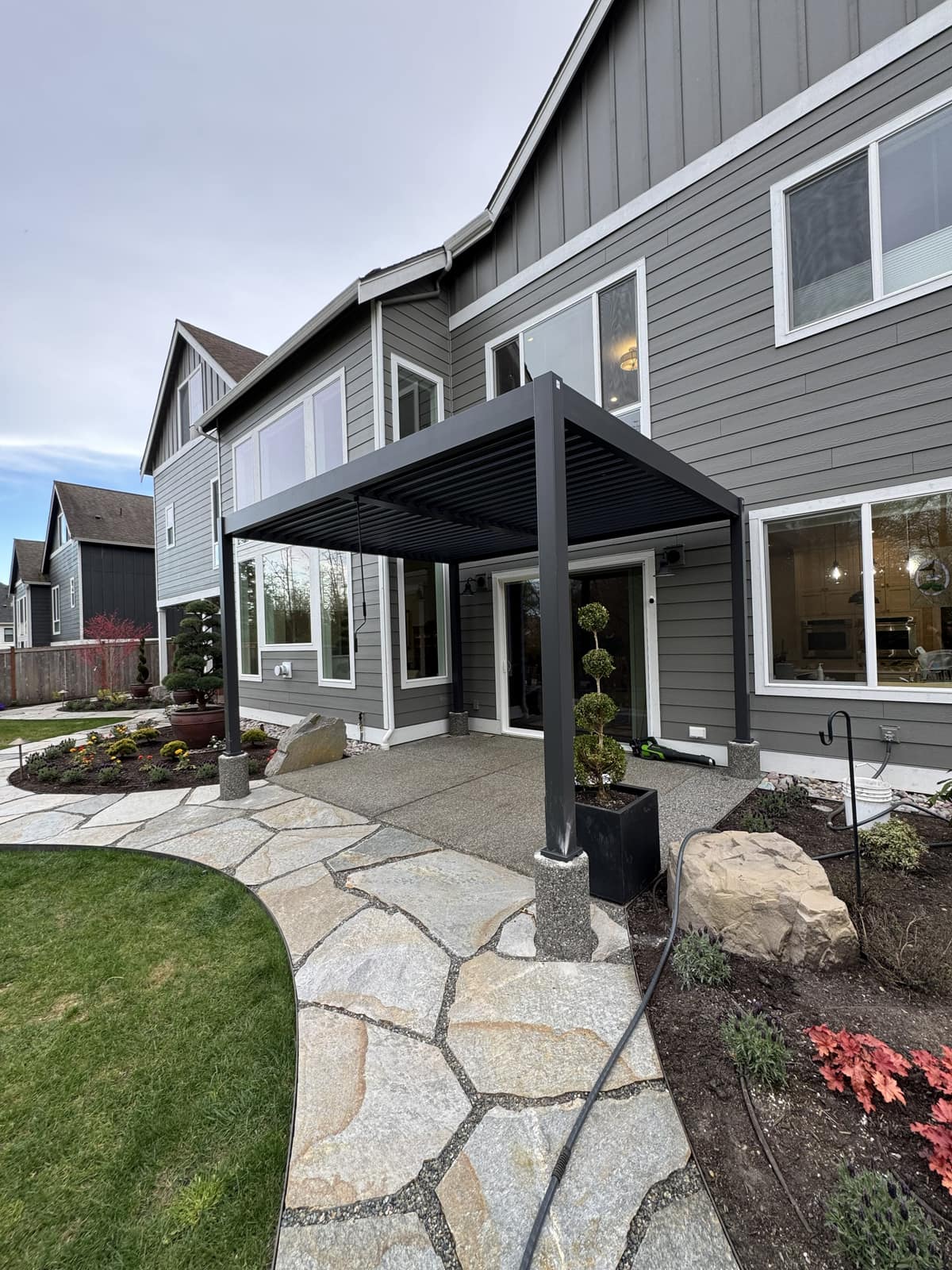 Black louvered pergola over a flagstone patio with topiary beside a gray home