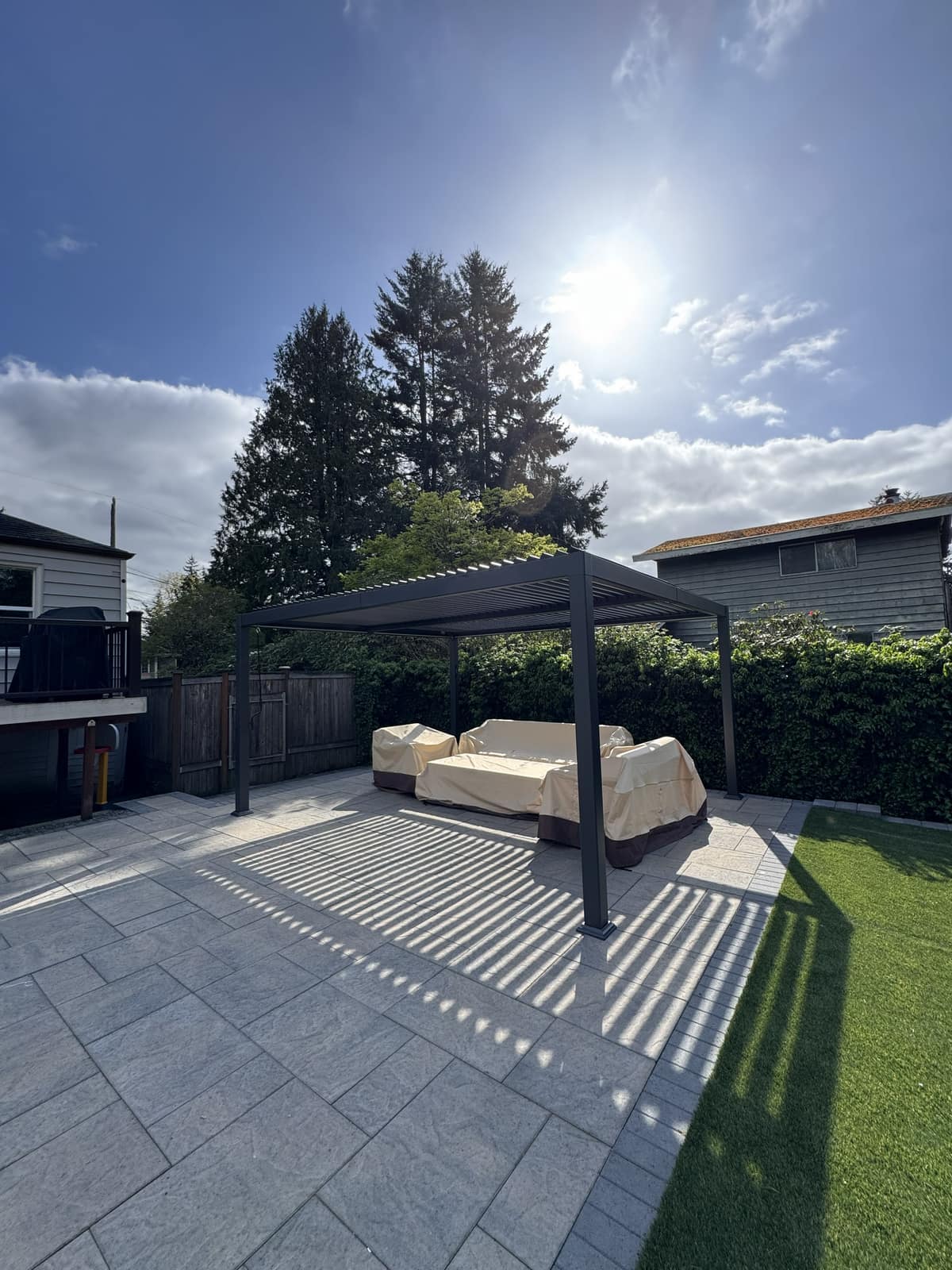 Black louvered pergola with covered furniture on a gray paver patio and blue sky