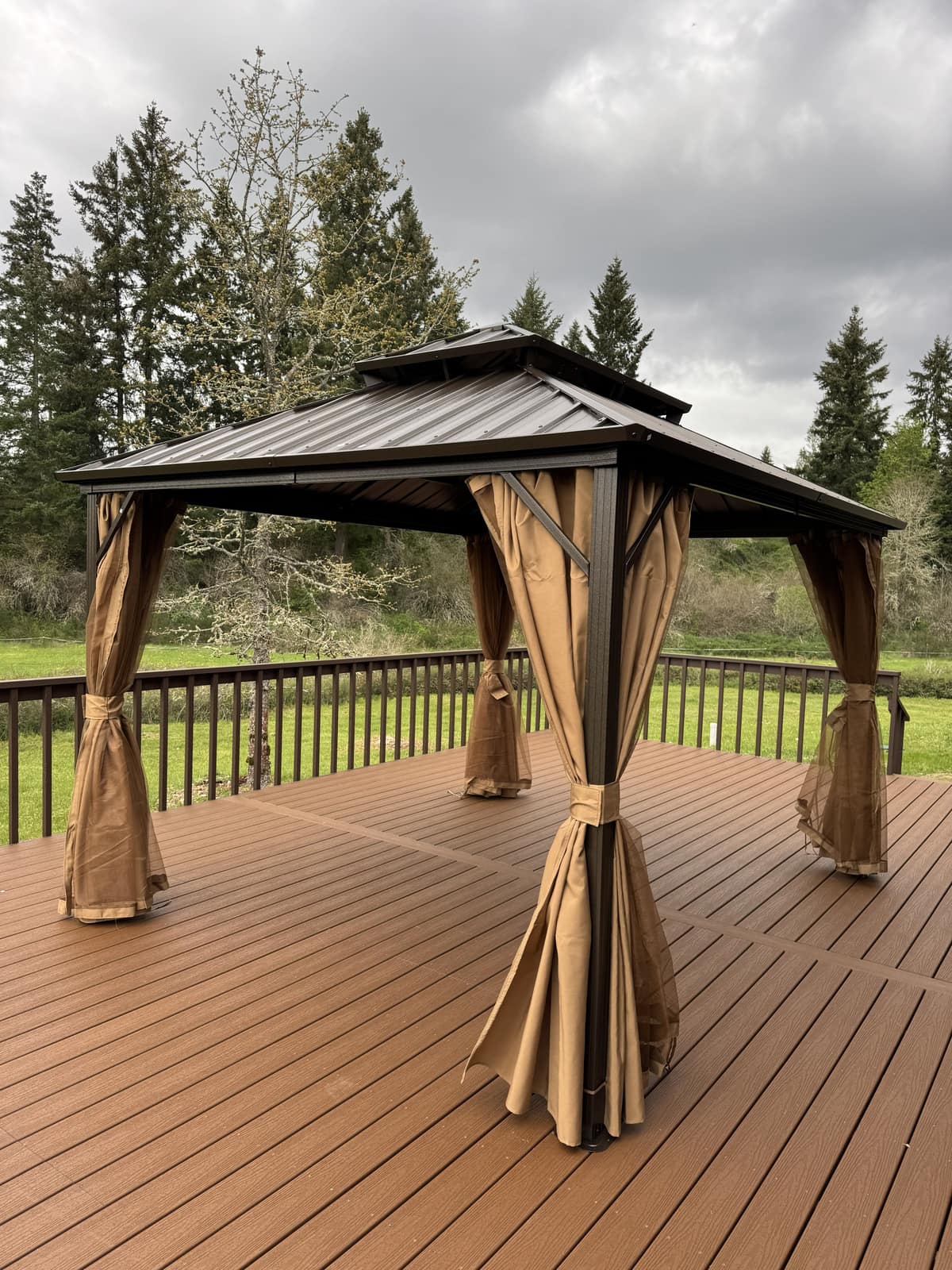 Bronze double-tier hardtop gazebo with curtains on a composite deck against a moody sky